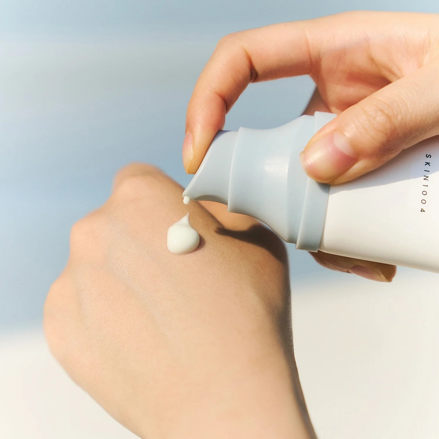 Primer plano de unas manos utilizando el dosificador del Hyalu‑Cica Water‑Fit Sun Serum de SKIN1004 para aplicar una gota de crema blanca en el dorso. La escena muestra la textura ligera tipo sérum del protector solar con ácido hialurónico y Centella Asiática que se absorbe rápido y no deja residuo blanco ni sensación grasa