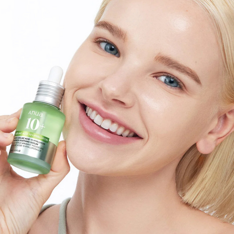 Retrato de una mujer rubia de piel clara, sonriendo y sujetando cerca de su mejilla un frasco de suero Anua 10+ con gotero, sobre fondo blanco.