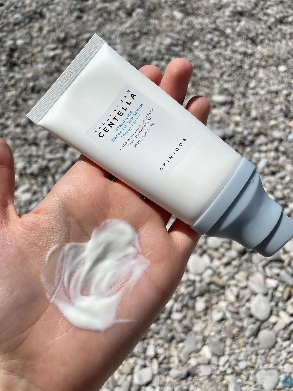 Mano sosteniendo el tubo del Hyalu‑Cica Water‑Fit Sun Serum de SKIN1004 sobre un lecho de piedras grises, con una cantidad generosa de crema blanca extendida en la palma. La fotografía destaca la textura cremosa y ligera del protector solar con ácido hialurónico y Centella Asiática de Madagascar, que proporciona hidratación y calma sin dejar manchas ni sensación grasa