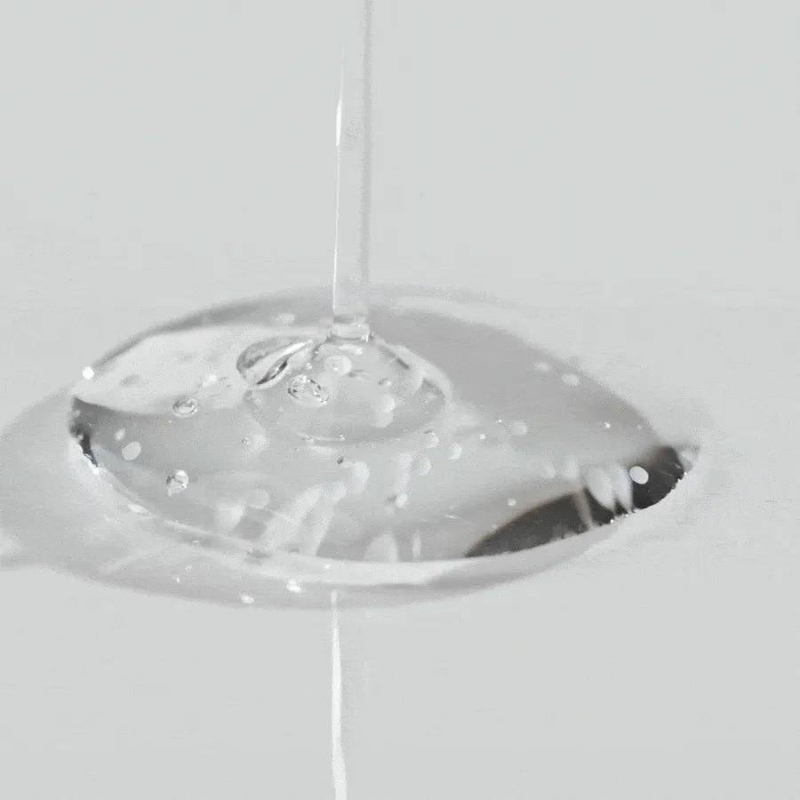 Primer plano de una gota transparente del sérum Tone Brightening Capsule Ampoule vertida en una superficie. Se observan microcápsulas suspendidas que contienen Madewhite™ encapsulado, niacinamida y ácido tranexámico para tratar manchas y unificar el tono