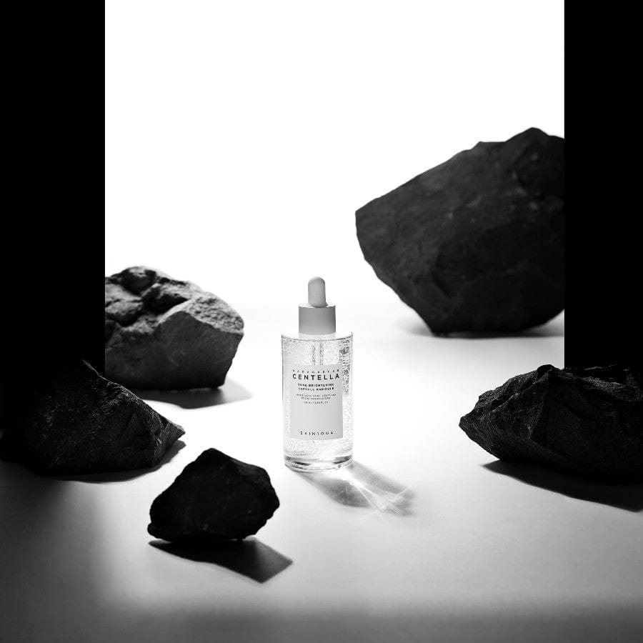Ampolla de SKIN1004 acompañada de rocas negras, mostrando las cápsulas brillantes del sérum iluminador. La imagen comunica la acción calmante y correctora de manchas del sérum, formulado con Centella Asiática de Madagascar, niacinamida y ácido tranexámico