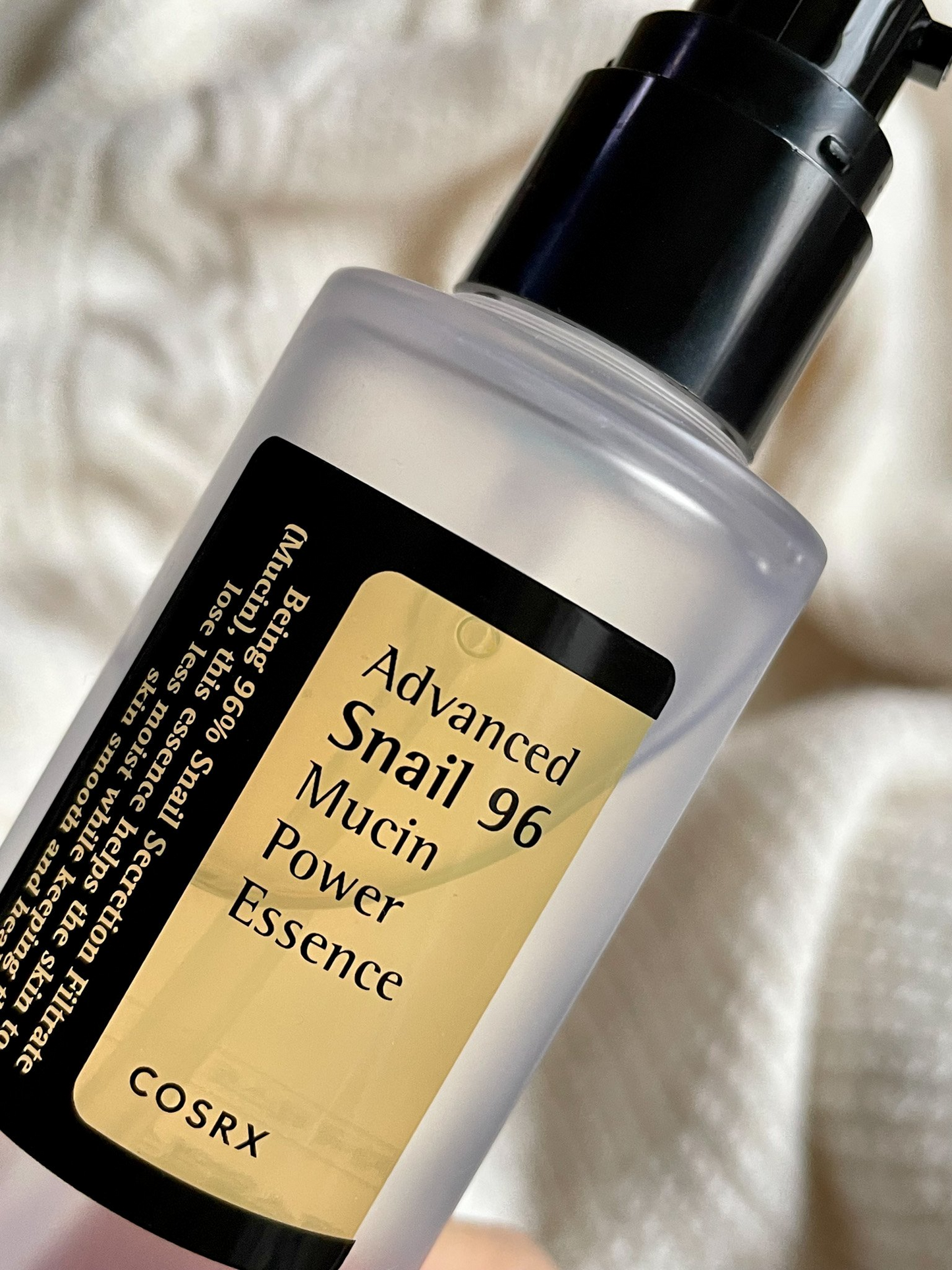 Botella transparente con etiqueta dorada y negra de COSRX Advanced Snail 96 Mucin Power Essence sobre fondo de tela blanca arrugada, en ángulo cercano.