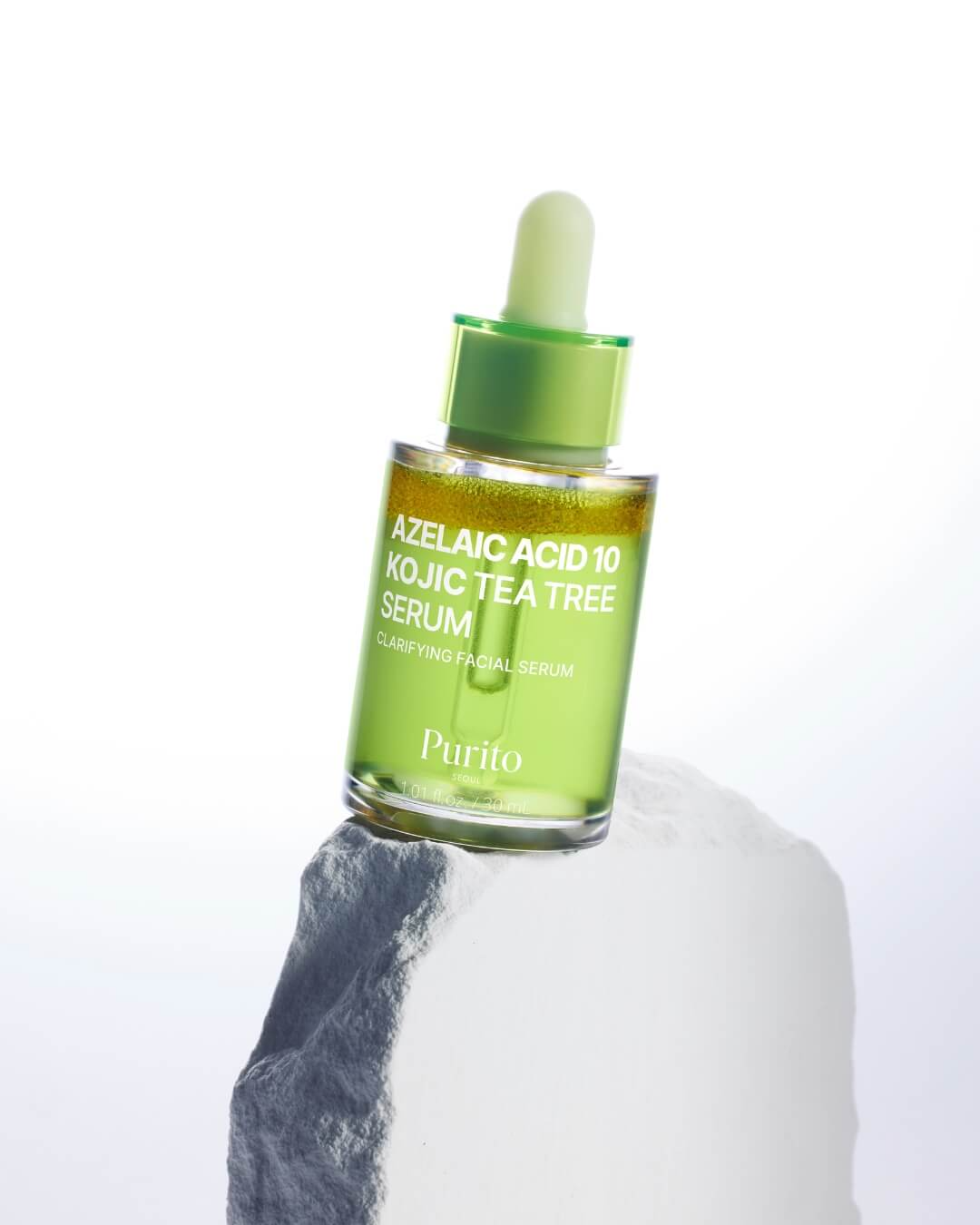 Frasco verde transparente del Purito Azelaic Acid 10 Kojic Tea Tree Serum sobre una roca blanca, iluminado con fondo blanco suave.
