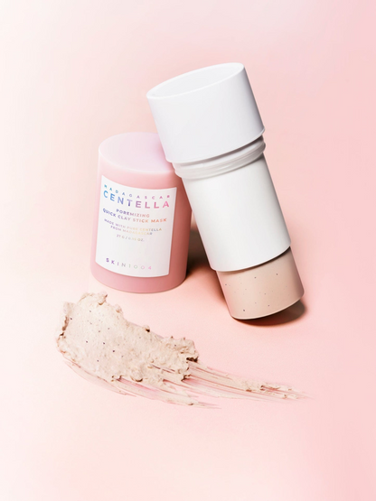 Alt-text:
Stick de mascarilla Skin1004 Madagascar Centella Poremizing Quick Clay Stick Mask con la barra abierta y textura beige extendida, fondo rosa degradado.