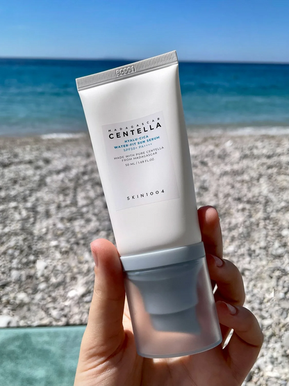Mano sosteniendo el tubo del Hyalu‑Cica Water‑Fit Sun Serum de SKIN1004 frente a una playa de guijarros y mar azul. El envase con base azul y etiqueta de Centella de Madagascar se resalta, sugiriendo su fórmula refrescante con ácido hialurónico y Centella Asiática que protege e hidrata la piel con SPF50+ PA++++ sin dejar sensación pesada