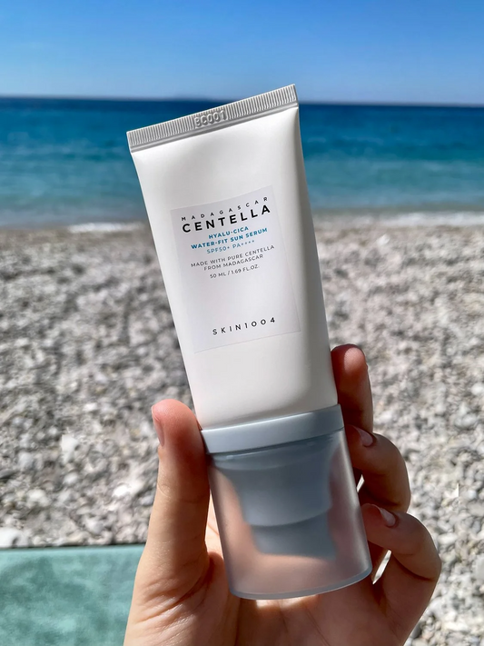 Mano sosteniendo el tubo del Hyalu‑Cica Water‑Fit Sun Serum de SKIN1004 frente a una playa de guijarros y mar azul. El envase con base azul y etiqueta de Centella de Madagascar se resalta, sugiriendo su fórmula refrescante con ácido hialurónico y Centella Asiática que protege e hidrata la piel con SPF50+ PA++++ sin dejar sensación pesada
