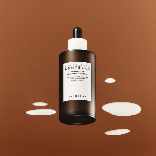 Ampolla Probio‑Cica Intensive de SKIN1004 con etiqueta claramente visible sobre fondo marrón y rodeada de manchas cremosas, destacando la textura lechosa y calmante del suero con extracto de Centella de Madagascar