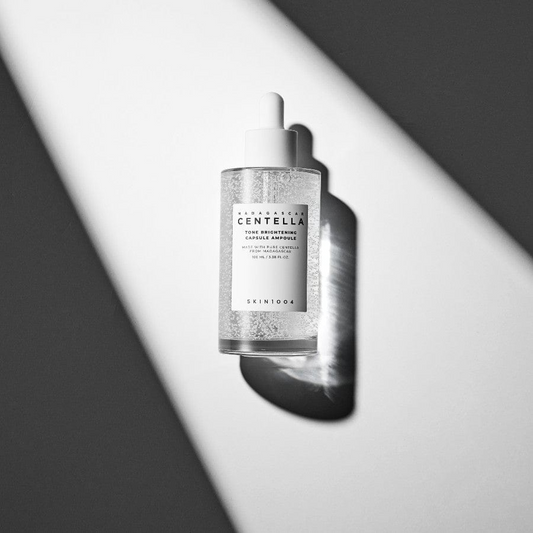 Ampolla de SKIN1004 iluminada por una banda diagonal de luz que proyecta sombras dramáticas. Se aprecian las microcápsulas del sérum con niacinamida, ácido tranexámico y Madewhite™ encapsulado para unificar e iluminar la piel