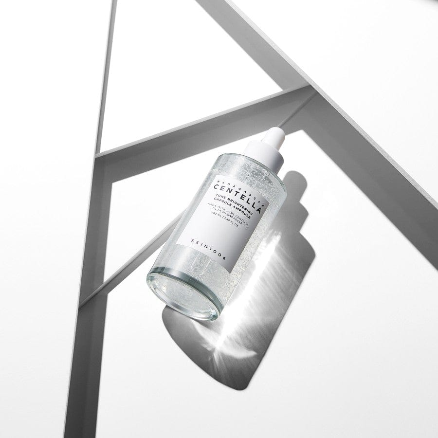 Botella del sérum unificador de tono inclinada sobre una estructura geométrica blanca con líneas y sombras, resaltando la ligereza del producto y su fórmula con Centella Asiática, niacinamida y ácido tranexámico que mejora la luminosidad y corrige manchas